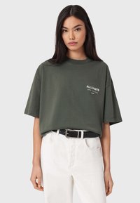 Oliivinvihreä oversized t-paita lyhyillä hihoilla, jossa valkoinen "ALLSAINTS"-logo vasemmassa rinnassa, yhdistetty korkeavyötäröisiin valkoisiin housuihin.