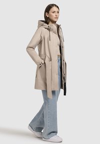 Beige waterdichte parka jas met een capuchon, drukknoopsluiting, verstelbare trekkoorden en grote zijzakken, gecombineerd met lichtblauwe jeans.