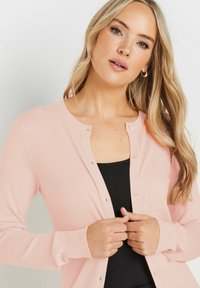 Cardigan rosa chiaro con scollatura rotonda, maniche lunghe e chiusura con bottoni frontali. Indossato sopra un top nero con texture a costine.