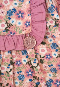 Květinová látka s růžovým volánem, zdobená malými modro-bílými květy. Z švu visí přívěsek z růžového zlata, který přidává texturu.