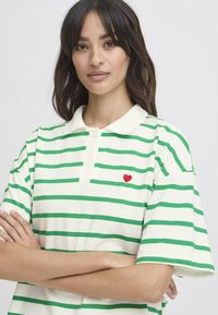 Kurzärmliges Polohemd in Creme mit grünen horizontalen Streifen. Es verfügt über einen Kragen und ein kleines rotes Herzsymbol auf der linken Seite.
