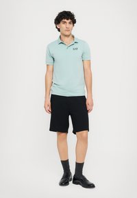 Svetlomodrá polo košeľa s krátkymi rukávmi a čiernym logom, kombinovaná s čiernymi šortkami. Model má na sebe aj šedé ponožky a čierne topánky.