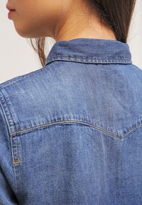 Chemise en jean avec un col classique, de couleur bleue, coutures apparentes et texture lisse, mettant en valeur un design arrière ajusté.