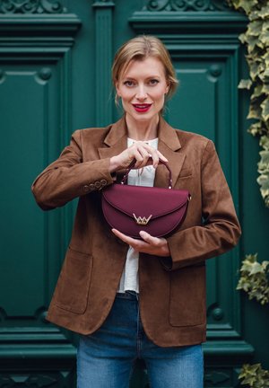 Burgunderfarbene Handtasche mit geschwungener Form, strukturierter Oberfläche und goldfarbener Kronen-Akzent. Von einer Person in einem braunen Wildlederblazer vor einem grünen Hintergrund gehalten.