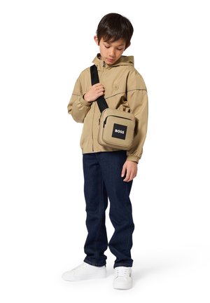 Chlapec má na sebe béžovú bundu s kapucňou, tmavomodré rifle, biele tenisky a béžovú tašku cez rameno s logom „BOSS“ na prednom vrecku.
