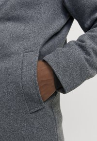 Main dans la poche d'un manteau en laine gris avec le poignet d'une manche intérieure noire visible sur un bras à la peau foncée.