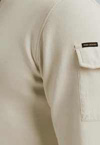 Top de algodón beige de manga larga con un bolsillo de parche en el hombro, que presenta la marca PME Legend en una etiqueta negra. Tejido de textura suave.