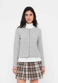 Γκρι ζακέτα με φερμουάρ και δύο μπροστινές τσέπες, κατασκευασμένη από υφασμάτινο με υφή. Φορεμένη πάνω από λευκό ζιβάγκο, συνδυασμένη με καρό φούστα.