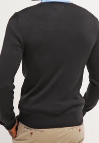 Pull noir à manches longues avec un col rond, une texture lisse et une coupe ajustée, porté sur une chemise bleu clair à col.