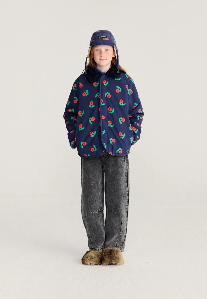 Enfant portant une veste fleurie marine et un chapeau assorti, un jean gris et des chaussons marron pelucheux, debout devant un fond uni.