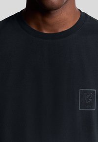 Zwarte katoenen T-shirt met een ronde hals en korte mouwen, voorzien van een subtiele grijze grafische logo-patch op de linkerkant van de borst.