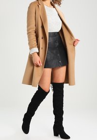 Cappotto di cammello, maglione grigio a maglia, mini gonna in pelle nera con dettagli a bottoni e stivali neri sopra il ginocchio con lacci.