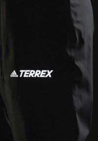 Pantaloni sportivi neri realizzati in tessuto morbido e leggero. Presentano una tasca con zip e un logo "TERREX" riflettente bianco con l'emblema Adidas.