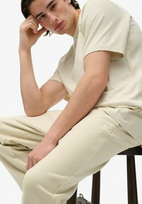 Beige katoenen t-shirt en bijpassende broek, met een relaxte pasvorm en subtiele merklogo op de broekspijp, zittend op een houten kruk.