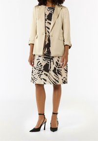 Blazer beige con mangas remangadas sobre un vestido de longitud a la rodilla con un patrón abstracto en negro y beige. Zapatos de tacón negros con correas en los tobillos.