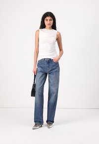 White sleeveless top with gathered texture, paired with high-waisted, loose-fit denim jeans. Black handbag and metallic shoes complete the look.