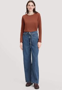 Vrouw staat gekleed in een roestkleurig langmouwtop, hoge taille wijde spijkerbroeken met een gestrikte riem en donkerbruine schoenen, op een lichte achtergrond.