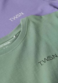 Deux T-shirts : l'un vert avec le texte noir "T.W.S.N.", l'autre violet avec le texte blanc "T.W.S.N.". En coton, col rond, texture lisse.