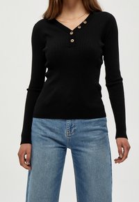 Svart ribbad långärmad tröja med V-ringning och fyra guldknappar på knappslån; kombinerad med ljusblå vida jeans.