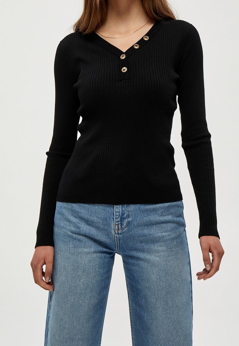 Svart ribbad långärmad tröja med V-ringning och fyra guldknappar på knappslån; kombinerad med ljusblå vida jeans.
