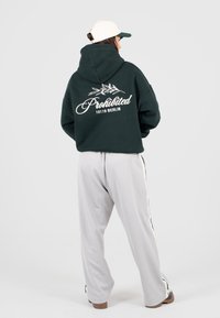 Persona che indossa un felpa verde scuro con il testo "Prohibited 10119 Berlin", pantaloni grigi chiari con stripe laterali, scarpe marroni e cappellino bianco rivolto di lato.