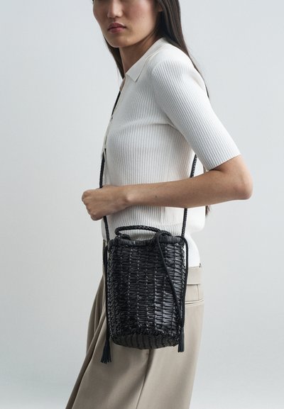 Μαύρη πλεκτή τσάντα χειρός με κυλινδρικό σχήμα, υφασμάτινη επιφάνεια και πλεγμένο λουρί ώμου. Διαθέτει κρόσσια στην κλειστή επένδυση.