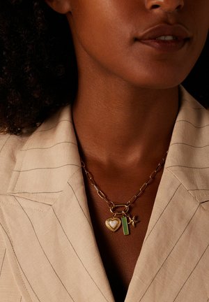 Close-up of woman wearing beige pinstripe blazer and gold chain necklace with heart, green rectangle, and star-shaped charms.