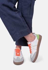 Białe trampki z szarymi zamszowymi akcentami, pomarańczowymi sznurówkami i zielonym szczegółem na pięcie. Gumowa podeszwa i perforowany design zapewniający oddychalność.