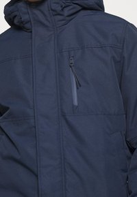Chaqueta azul marino hecha de material duradero, con un bolsillo en el pecho con cremallera, capucha ajustable y dos bolsillos laterales, de textura suave.