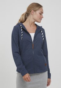 Mornarska zip-up kapuca, izdelana iz mehke tkanine, z naramnicami v črtastem vzorcu, z zadrgo in majhnim rjavim usnjenim logotipom.