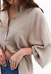 Camicetta beige, a trama, con colletto e maniche ampie, caratterizzata da sottili linee verticali. Abbinata a jeans blu.
