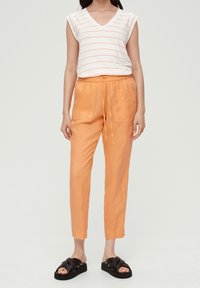 Witte V-hals t-shirt met oranje strepen, gecombineerd met lichte oranje linnen broek met een trekkoord in de taille en zijzakken. Zwarte schuivende sandalen.