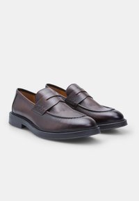 Mocassins en cuir marron à la finition lisse, avec un design cousu et une semelle en caoutchouc noir légèrement surélevée. Style classique à enfiler.