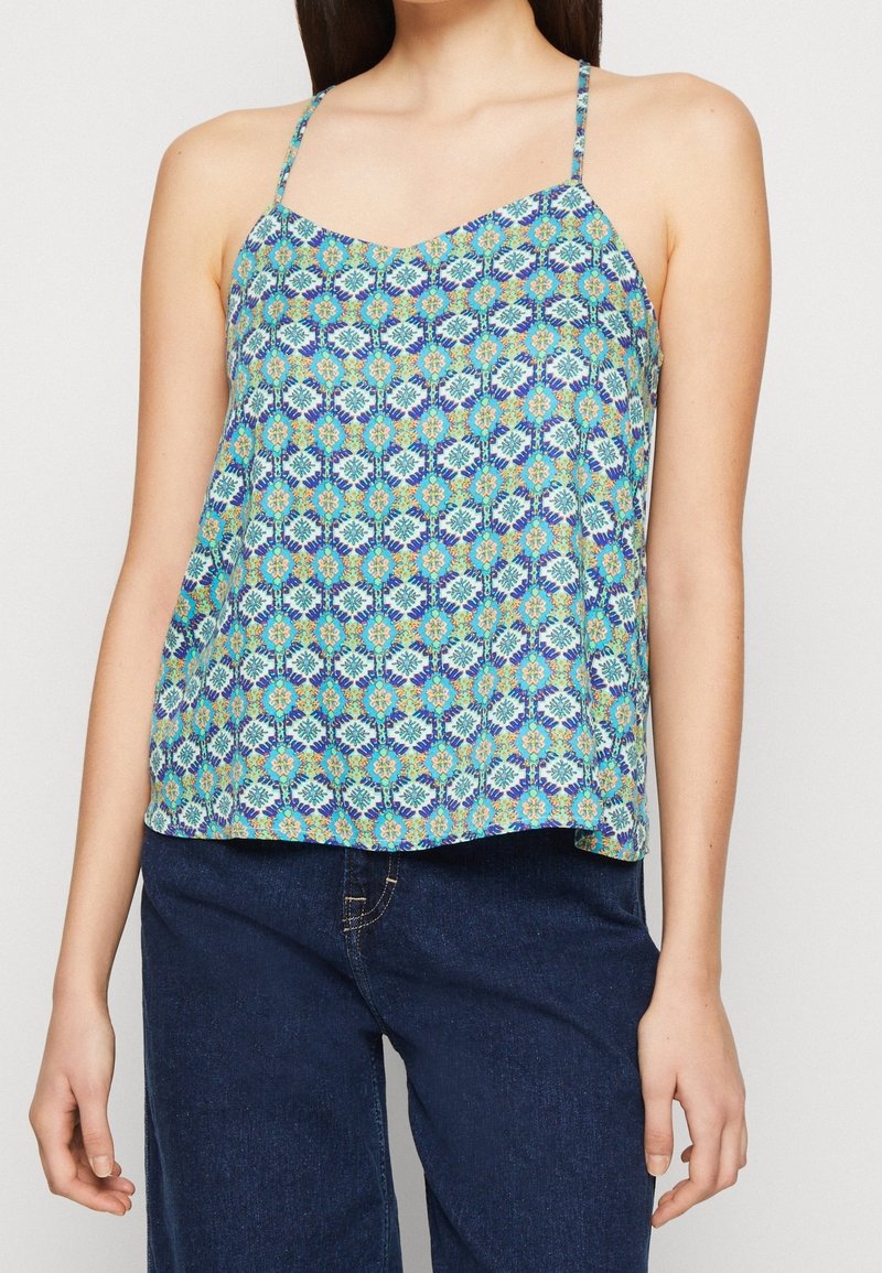 Femme portant un caraco sans manches bleu à motif floral et un jean en denim bleu foncé devant un fond uni.