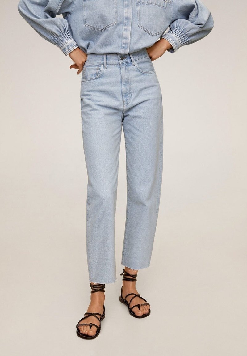 Personne portant un jean droit taille haute bleu clair, une veste en denim bleu clair, et des sandales plates noires à lanières avec des attaches à la cheville.