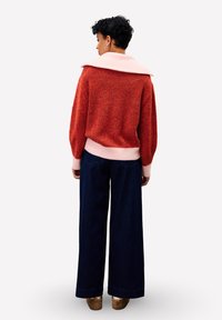 Sweater en maille côtelée rouge avec un col rose, associé à un jean large bleu foncé. La tenue présente un design décontracté et un tissu texturé.
