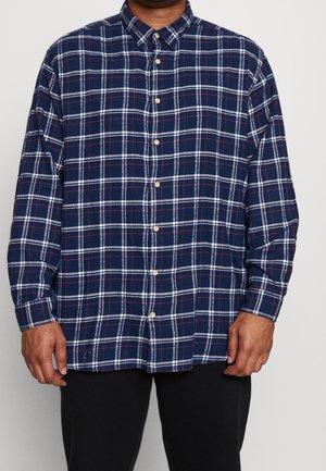 Homme portant une chemise bleu marine à manches longues avec un motif à carreaux blanc et rouge, associée à un pantalon noir sur un fond uni.