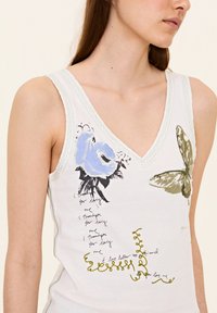 Witte katoenen tanktop met een V-hals, versierd met blauwe bloemen- en vlinderafdrukken, groene accenten en handgeschreven tekstdetails.
