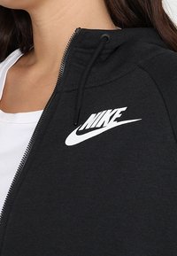 Svart zip-up hoodie gjord av bomullsblandning, med en vit Nike-logotyp på bröstet och justerbara dragsnören i huvan.