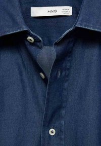 Tummansininen denimpaita, jossa on napituskaulus, valkoiset napit ja tagi, joka osoittaa koon M ja merkin MNG. Silkkinen pinta.