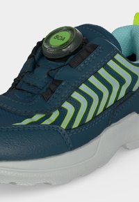 Gros plan sur une basket bleue avec des bandes vert fluo, une fermeture à molette BOA, une doublure intérieure bleu clair et une semelle blanche épaisse.