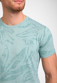 Šviesiai mėlyna marškinė su tekstūruotu lapų raštu ir mažo logotipo ženkleliu ant krūtinės. Trumpomis rankovėmis, apvaliu kaklu ir minkšta medžiaga.