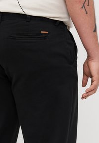 Black pants with a smooth texture, fitted style, and a back pocket featuring a branded leather label. Hand resting on the side.