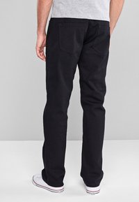 Schwarze Baumwollhosen mit geradem Bein, zwei Gesäßtaschen und dezenten Nähdetails. Getragen mit weißen Schuhen.