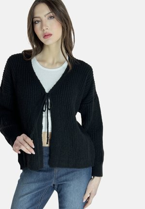 Schwarze gestrickte Strickjacke aus strukturiertem Stoff, mit offenem Vorderteil und Kordelzug. Getragen über einem weißen Oberteil und kombiniert mit blauen Jeans.