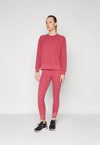 Felpa color rosa con maniche raglan, abbinata a leggings coordinati. Scarpe da ginnastica nere con accenti rosa. Tessuto liscio e aderente.