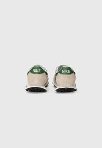 Tênis Nike com parte superior em camurça bege, detalhes em verde no calcanhar, texto do logotipo e sola de borracha preta com padrões de textura.