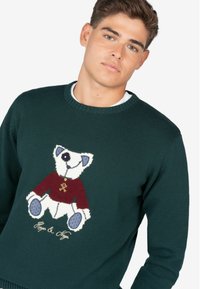Suéter verde de punto con un diseño de oso que lleva una camiseta roja. Los detalles notables incluyen ojos y patas bordados, además de un texto debajo del diseño.
