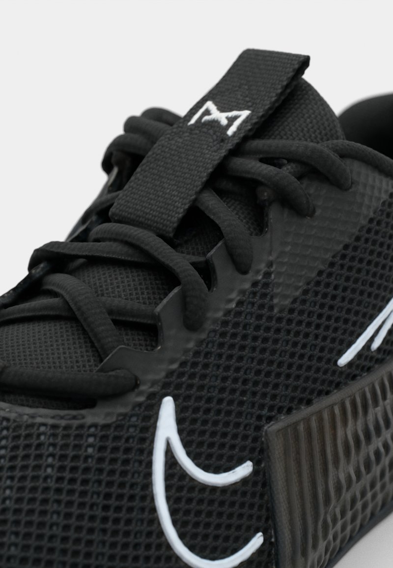 Chaussure de sport noire avec une tige en mesh texturé, des accents blancs contrastants et des lacets noirs plats. Présente une étiquette de marque sur la languette.
