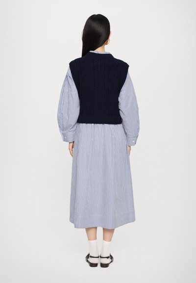 Marineblau gestrickte Weste mit strukturierten Rautenmuster über einem blau-weißen gestreiften langen Kleid. Das Outfit verfügt über Puffärmel und einen knöchellangen Rock.